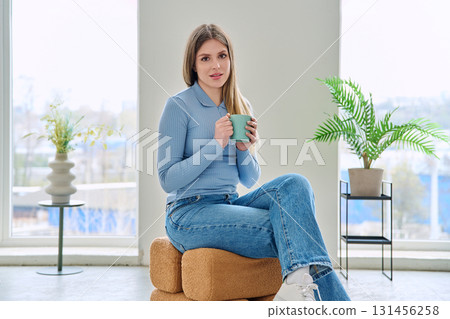Portrait of relaxed beautiful young woman sitting at home on chair with cup 131456258