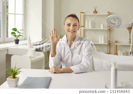 Happy woman beautician sitting at table, starting online skincare consultation and waving hello 131456843