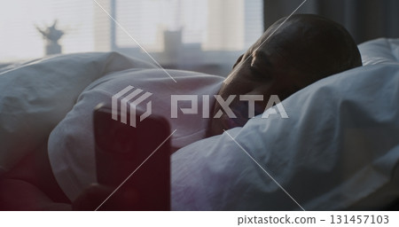 Man in Bed, Resting Head on White Pillow, Scrolling Through Social Media Content in Bright Sunlight 131457103
