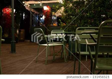 Cozy, dimly lit cafe terrace with metal green chairs and table, surrounded by plants and lights, evokes peaceful, intimate atmosphere perfect for relaxation or quiet conversations under night sky 131458323