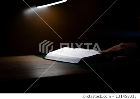 Person reading book at dimly lit desk, with light casting soft glow on pages. Evokes sense of quiet concentration, introspection, and peaceful atmosphere ideal for studying or reading Person reading book at dimly lit desk, with light casting soft glow on pages. Evokes sense of quiet concentration, introspection, and peaceful atmosphere ideal for studying or reading 131458333