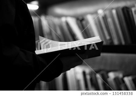 Person holds book in their hands, with pages turned, surrounded by bookshelves in soft focus. Black and white tone emphasizes calm, thoughtful moment, capturing essence of quiet reflection Person holds book in their hands, with pages turned, surrounded by bookshelves in soft focus. Black and white tone emphasizes calm, thoughtful moment, capturing essence of quiet reflection 131458338