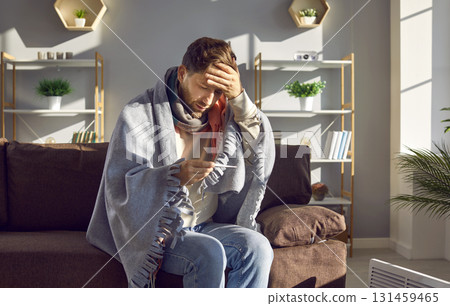 Man feeling cold sitting on sofa at home measuring temperature with thermometer Man feeling cold sitting on sofa at home measuring temperature with thermometer 131459465
