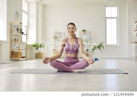 Young woman sitting on workout mat at home, doing yoga, meditating, and relaxing Young woman sitting on workout mat at home, doing yoga, meditating, and relaxing 131459494