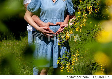 Pregnant couple forming heart shape on belly in blooming garden 131460094