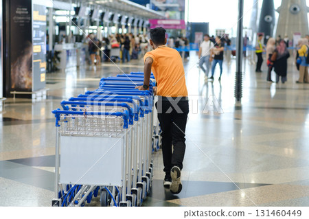 Man pushes luggage carts through busy airport terminal during daytime as travelers move around and check in for their flights Man pushes luggage carts through busy airport terminal during daytime as travelers move around and check in for their flights 131460449