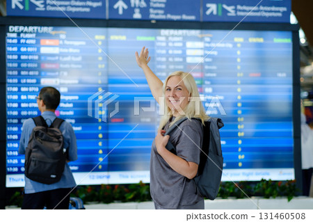 Woman with blonde hair smiles and points at departure board while standing at busy airport terminal in the afternoon, preparing for her upcoming flight departing soon 131460508