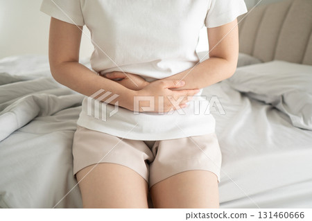 Young female suffering form stomach ache while sitting on a bed at home. Causes of abdominal pain include menstruation pain, gastritis, stomach ulcer, food poisoning, diarrhea or IBS. Young female suffering form stomach ache while sitting on a bed at home. Causes of abdominal pain include menstruation pain, gastritis, stomach ulcer, food poisoning, diarrhea or IBS. 131460666