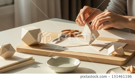 Person crafting geometric wooden shapes on natural wood cutting board with scissors and white paper materials in sunlit workspace 131461196