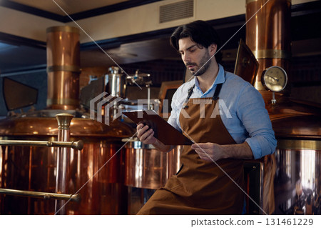 A man wearing an apron is studying a clipboard in a brewery setting 131461229