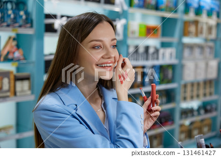 A woman is carefully applying lipstick to her lips while in a store nearby 131461427