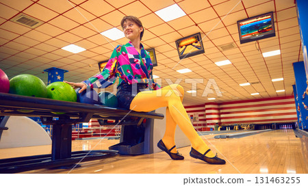 Woman sitting on bowling rack with colorful balls and playful smile 131463255