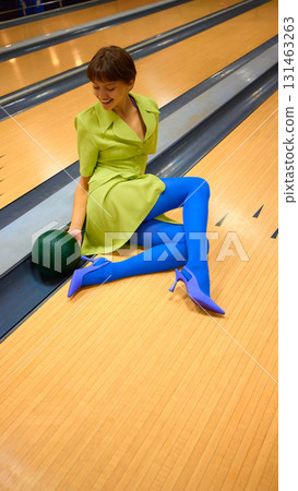 Woman in green blazer and blue tights sitting on bowling lane with ball Woman in green blazer and blue tights sitting on bowling lane with ball 131463263