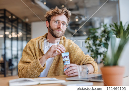 Man sitting at coworking desk eating plant based snack package labeled high protein zero sugar Man sitting at coworking desk eating plant based snack package labeled high protein zero sugar 131464103
