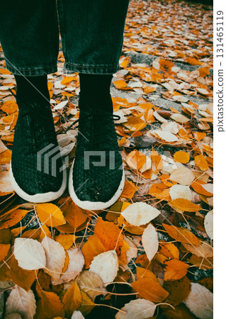Black sneakers on autumn leaves, a perfect fall scene with colorful foliage. A person's feet in black sneakers stand on fallen autumn leaves. Jeans and socks are also visible. Black sneakers on autumn leaves, a perfect fall scene with colorful foliage. A person's feet in black sneakers stand on fallen autumn leaves. Jeans and socks are also visible. 131465119