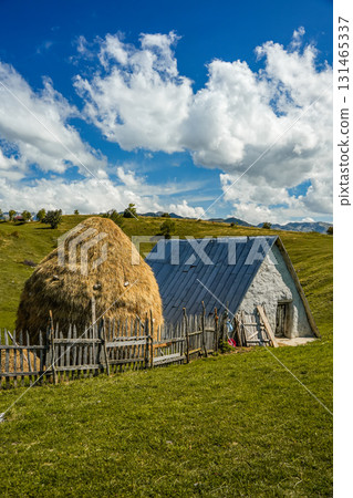 Scenic view of a single clay house with a haystack nearby under white clouds, showcasing traditional rural landscape, ideal for travel, lifestyle, and nature projects. 131465337