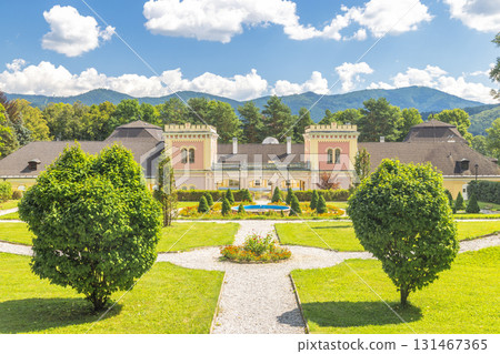 Hodkovce Mansion in Zehra village in Slovakia, Europe. Stately manor house nestled in a lush green park with a mountain backdrop under a clear blue sky filled with fluffy white clouds. 131467365