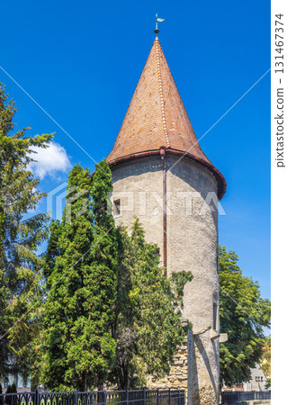 Bardejov town in North-Eastern Slovakia, Europe. Stone tower rises, topped with a conical tile roof, framed by lush green trees against a bright blue sky. Historical architecture. Bardejov town in North-Eastern Slovakia, Europe. Stone tower rises, topped with a conical tile roof, framed by lush green trees against a bright blue sky. Historical architecture. 131467374