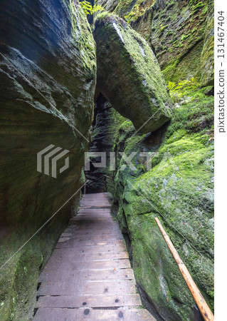 Adrspach-Teplice Rocks, sandstone formations in Hradec Kralove Region in the Czech Republic, Europe. A narrow wooden pathway through mossy rock formations, illuminated by light at the distant end. 131467404