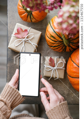 Top view of woman holds smartphone with blank white screen, surrounded by pumpkins, gifts wrapped in craft paper and autumn leaves. Ideal for displaying app concepts or holiday promotions Top view of woman holds smartphone with blank white screen, surrounded by pumpkins, gifts wrapped in craft paper and autumn leaves. Ideal for displaying app concepts or holiday promotions 131467550