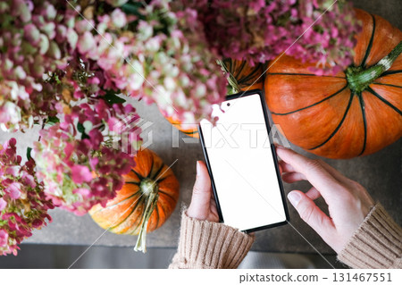 Woman holds a smartphone with blank white screen, surrounded by pumpkins and autumn flowers. Cozy, celebratory mood of the season, perfect for showcasing mobile apps or holiday content Woman holds a smartphone with blank white screen, surrounded by pumpkins and autumn flowers. Cozy, celebratory mood of the season, perfect for showcasing mobile apps or holiday content 131467551