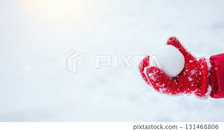 A gloved hand, covered in white snowflakes, holds a perfectly round snowball against a bright, soft white winter background with subtle sunlight. 131468806
