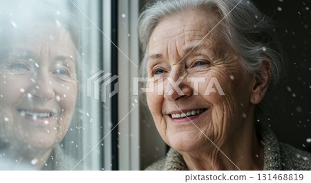 A heartwarming closeup of a serene elderly woman with grey hair and striking blue eyes, smiling while looking out a window at falling snowflakes, her reflection clearly visible. 131468819
