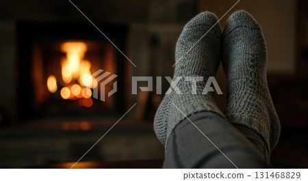 A person's feet wearing warm grey knitted socks are crossed and elevated, resting comfortably in front of a cozy, glowing fireplace with blurred orange flames providing a soft, intimate light. 131468829