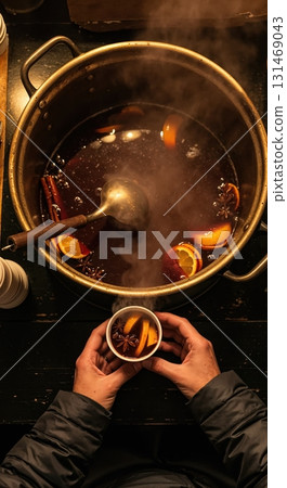 A top-down view of a steaming pot of mulled wine with orange slices, cinnamon sticks, and star anise, as hands in a dark jacket hold a cup of the warm beverage. 131469043