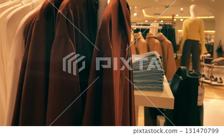 View of clothing racks, folded jeans, and mannequins inside a generic fashion retail store. Stylish contemporary interior with warm lighting and neatly arranged garments View of clothing racks, folded jeans, and mannequins inside a generic fashion retail store. Stylish contemporary interior with warm lighting and neatly arranged garments 131470799