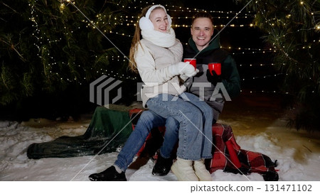 Winter evening warmth, young couple cuddling on blanket covered bench, holding steaming mugs, smiling amid gently falling snow and twinkling christmas lights Winter evening warmth, young couple cuddling on blanket covered bench, holding steaming mugs, smiling amid gently falling snow and twinkling christmas lights 131471102
