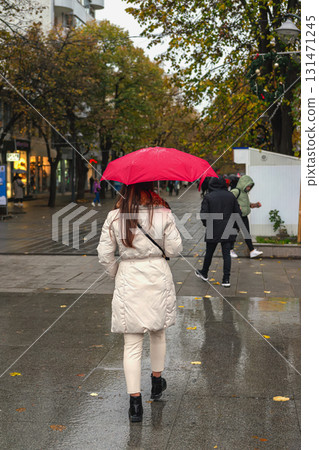 Woman in light coat holds a red umbrella while walking on a rainy autumn city street, rear view. Concept of autumn, rain, lifestyle in wet seasonal weather 131471245