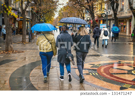 Three women walking under umbrellas on wet city street lined with autumn trees. Concept of autumn, rain, urban lifestyle in seasonal weather 131471247