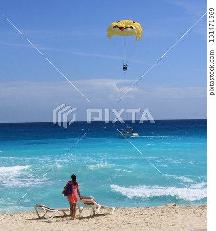 Woman in a red dress watches a bright yellow parasail lift tourists above the turquoise ocean 131471569
