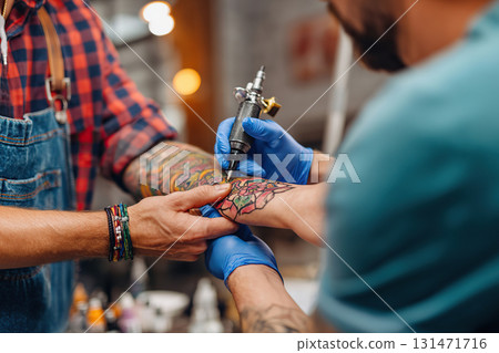 Tattoo artist inking a detailed floral design on a client's forearm in a bustling studio 131471716