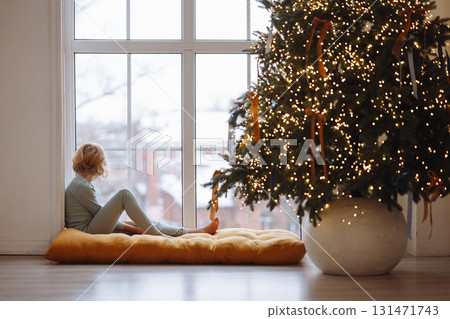 Girl sitting on cushion by window near decorated Christmas tree with lights in cozy room Girl sitting on cushion by window near decorated Christmas tree with lights in cozy room 131471743