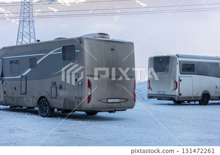 Two motorhomes stand parked on a snowy lot with dirt covering their rear sides. The frozen ground and misty mountain background highlight winter travel conditions Two motorhomes stand parked on a snowy lot with dirt covering their rear sides. The frozen ground and misty mountain background highlight winter travel conditions 131472261