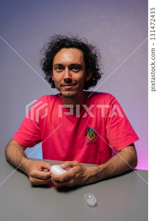 Vertical portrait of handsome young adult man with curly hair and pink t-shirt cutting fingernails using nail clipper sitting at gray table against purple background, smiling looking at camera. 131472435