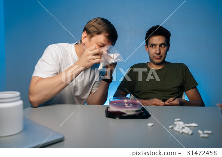Two young men sitting at table with birthday cake, one snorting powdered drug while other watches, illustrating risks of youth substance abuse. Concept of party atmosphere with substance abuse. Two young men sitting at table with birthday cake, one snorting powdered drug while other watches, illustrating risks of youth substance abuse. Concept of party atmosphere with substance abuse. 131472458