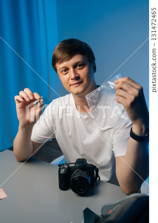 Vertical portrait of photographer male sitting at desk with camera and showing rolled up banknote and plastic bag with cocaine, depicting drug addiction issues. Concept of substance abuse. Vertical portrait of photographer male sitting at desk with camera and showing rolled up banknote and plastic bag with cocaine, depicting drug addiction issues. Concept of substance abuse. 131472465