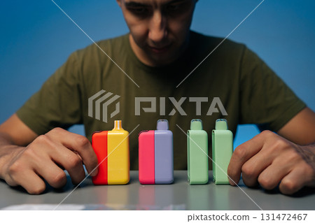 Blurred male holding and presenting various new heat-not-burn tobacco heating systems in vibrant colors on gray table, against isolated blue background. 131472467