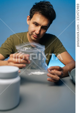 Vertical portrait of drug dealer male preparing dose of cocaine or methamphetamine from plastic bag, with white plastic jar and scales in blurred background. Concept of drug dealing or addiction. 131472469