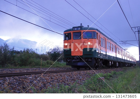 Shinano Railway 115 series train (Shonan color) running through Karuizawa on a cloudy day, watched over by Mount Asama. Photo taken on September 27, 2025. Shinano Railway 115 series train (Shonan color) running through Karuizawa on a cloudy day, watched over by Mount Asama. Photo taken on September 27, 2025. 131473605