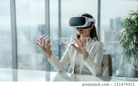 Woman in stylish white virtual reality headset gesturing with her hands sits at modern desk office, panoramic window overlooking cityscape. Woman in stylish white virtual reality headset gesturing with her hands sits at modern desk office, panoramic window overlooking cityscape. 131474351