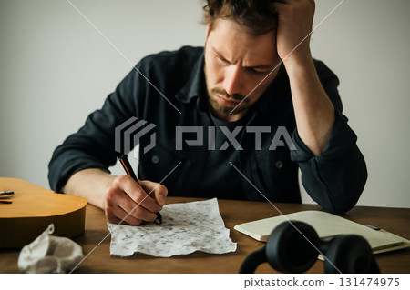 AI generated image of a musician deeply focused on writing lyrics on crumpled paper, showcasing the frustration and inspiration in a creative process surrounded by artistic clutter soft natural light. 131474975