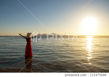 Young woman wearing long red dress and straw hat raising hands standing in sea water at the beach enjoying view of rising sun in early summer morning. 131475699