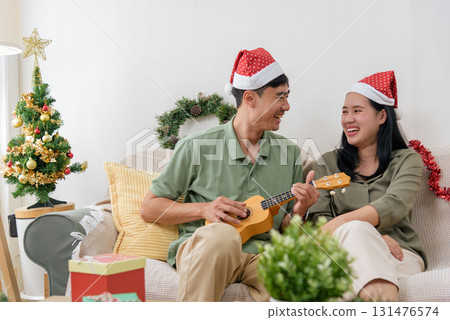 Christmas love, happy couple wears festive hats enjoying winter holiday at home, man plays ukulele for woman, celebrating special christmas time together surrounded by cheerful decorations. Christmas love, happy couple wears festive hats enjoying winter holiday at home, man plays ukulele for woman, celebrating special christmas time together surrounded by cheerful decorations. 131476574