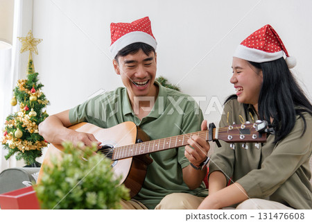 Christmas couple shares love, happy holiday winter moment with music as man plays guitar for woman wearing santa hats, enjoying cheerful festive christmas celebration at cozy home. Christmas couple shares love, happy holiday winter moment with music as man plays guitar for woman wearing santa hats, enjoying cheerful festive christmas celebration at cozy home. 131476608