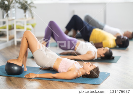 Multinational students lie on mat, put their foot on fitball and lift pelvis, raise and lower pelvis. Multinational students lie on mat, put their foot on fitball and lift pelvis, raise and lower pelvis. 131476687