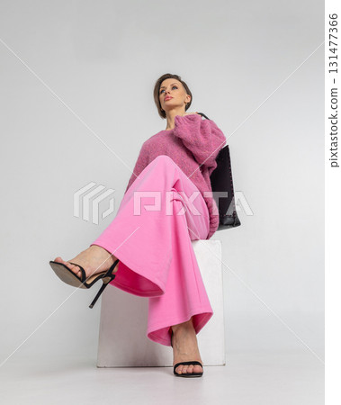 Portrait beautiful woman of model appearance. Girl in pink sweatshirt and trousers, black bag on grey background. Pretty female with trendy makeup posing in studio. Vertical photo. 131477366